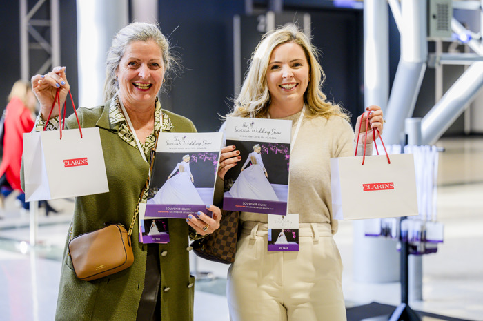 Two women smile holding a wedding show programme and a Clarins gift bag each 