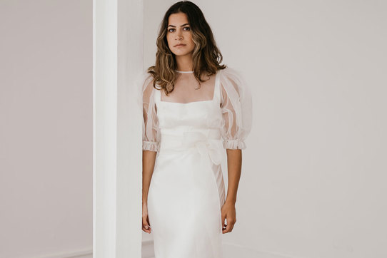 brunette woman wears a form-fitting silky whiten gown with organza puff sleeved top as an added layer