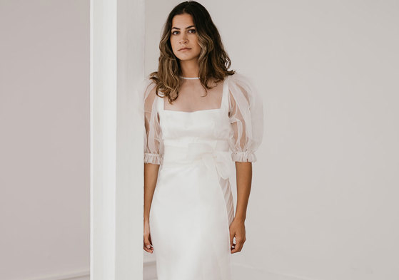 brunette woman wears a form-fitting silky whiten gown with organza puff sleeved top as an added layer