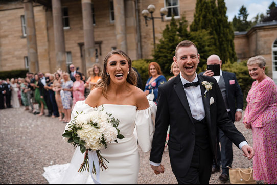 Bride and groom laugh as they leave their ceremony with guests clapping in background