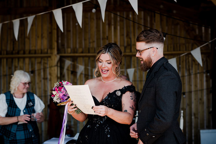A Bride And Groom Both Wearing Black Singing While Looking At A Sheet Of Paper Held By The Bride, Set Against A Wooden Shed Building Hanging With White Bunting