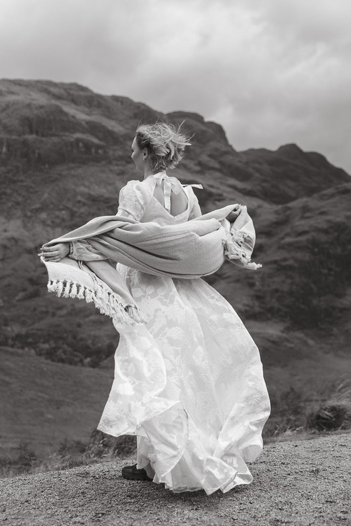 bride with shoulder length hair and floral embellished wedding dress holds a shawl across her arms, extending them outwards, facing the wind and view of nearby hills