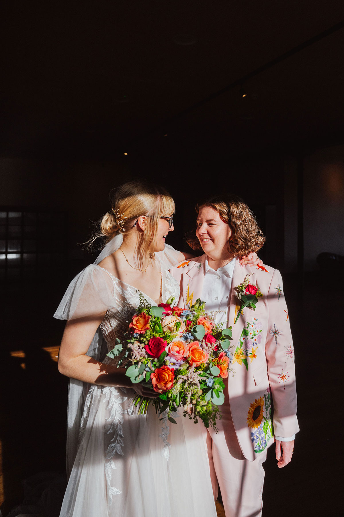 Two brides standing by a sunlit industrial window at the Engine Works, smiling at each other with a bright, colourful bouquet.