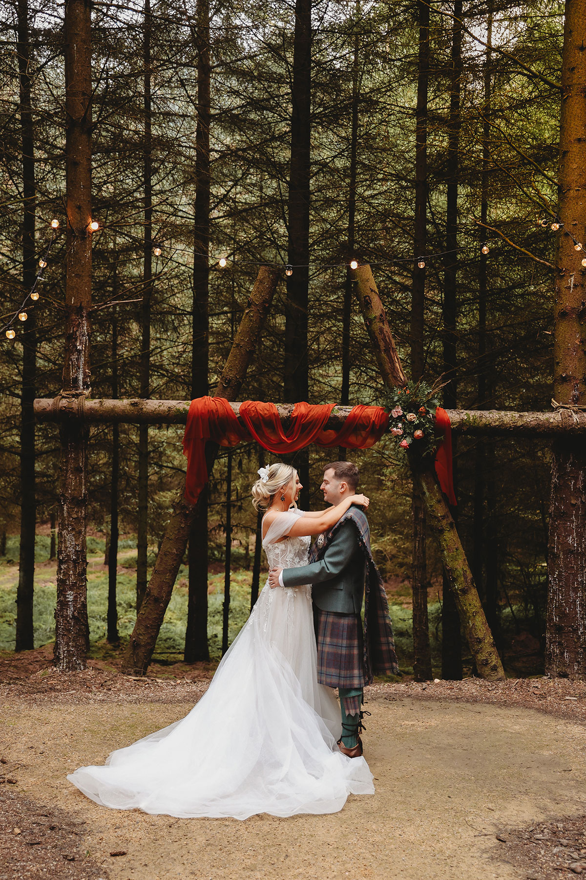 Bride and groom embracing beneath a rustic woodland ceremony arch decorated with red draping at a Scottish wedding