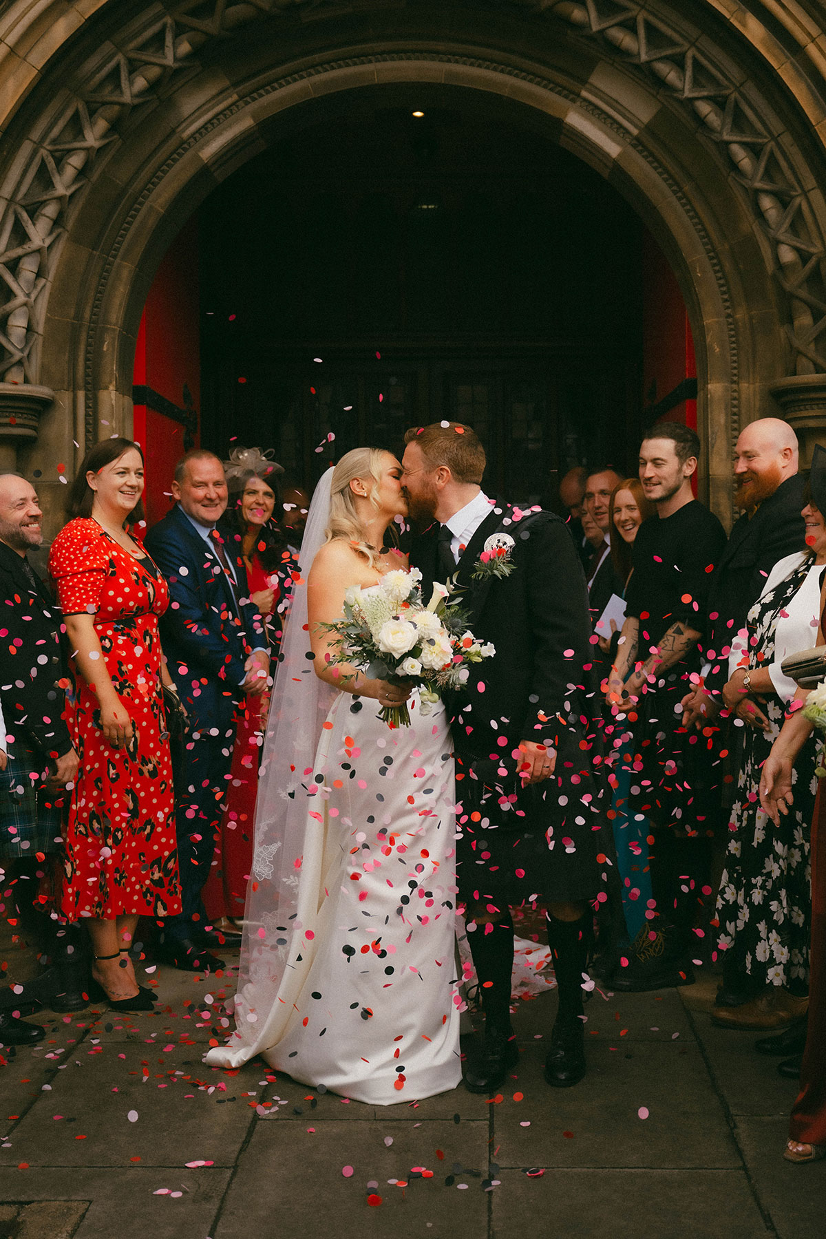newlyweds kissing outside church with red doors and confetti throw by guests
