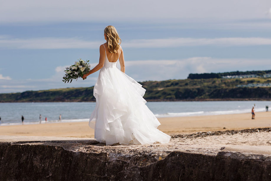 A bride at her Old Course Hotel wedding in a beautiful white gown gazes out at the sea from a sandy beach, holding a bouquet and contemplating the horizon
