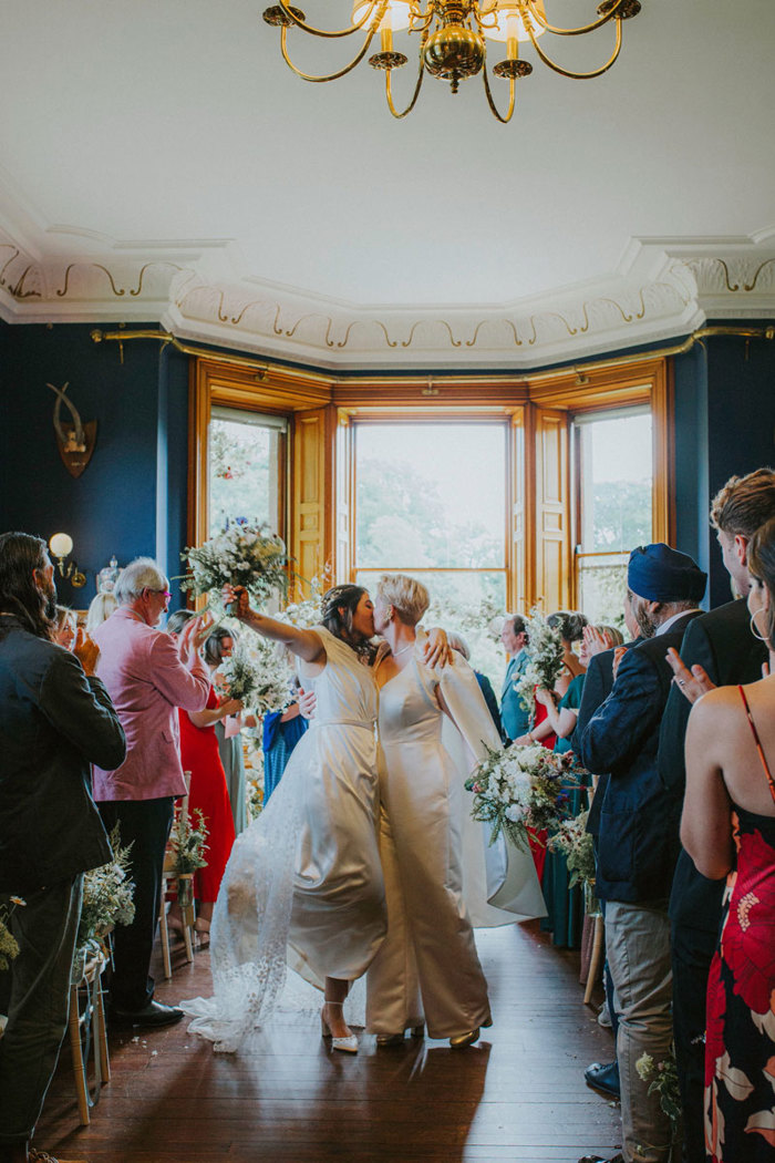 two brides kiss and hold bouquets in the air as guests cheer during wedding at cambo house