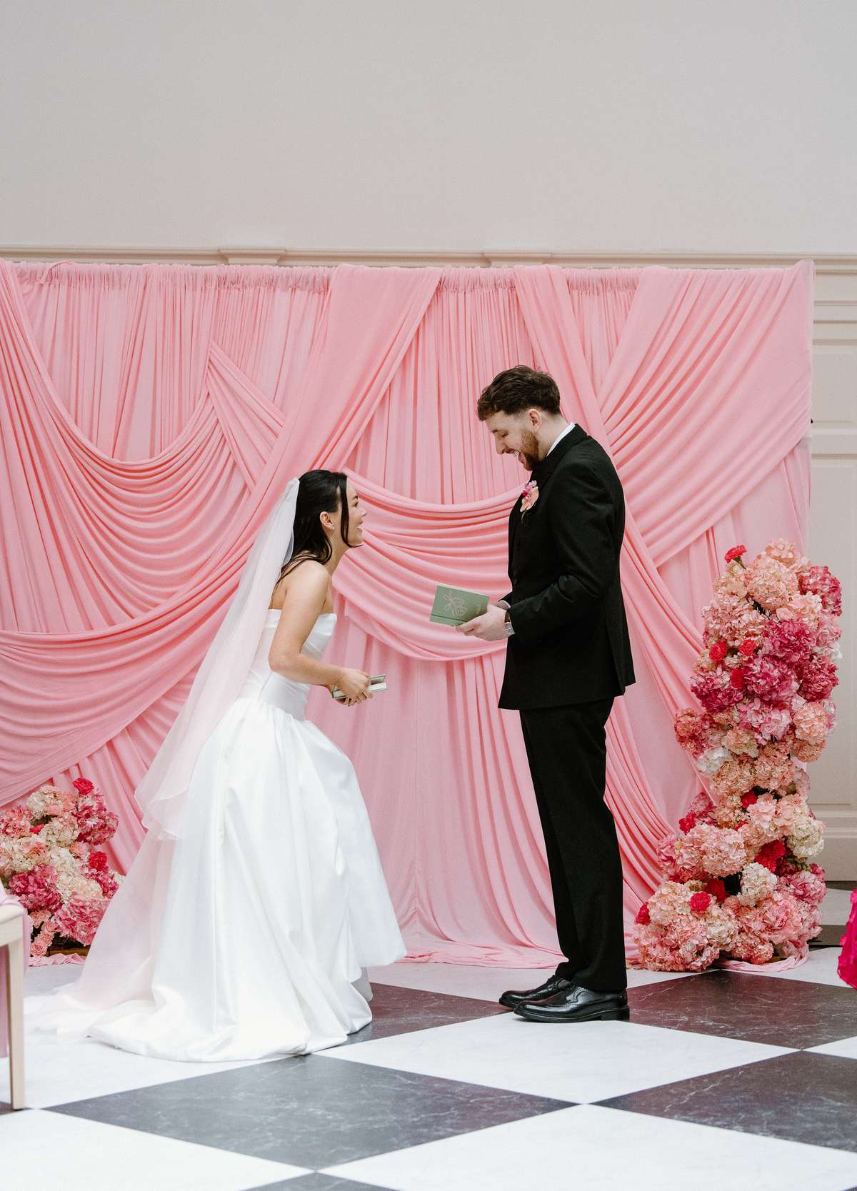 Bride and groom exchange personal wedding vows in front of a pink draped backdrop and colourful floral arrangements at The Collector’s Hall.
