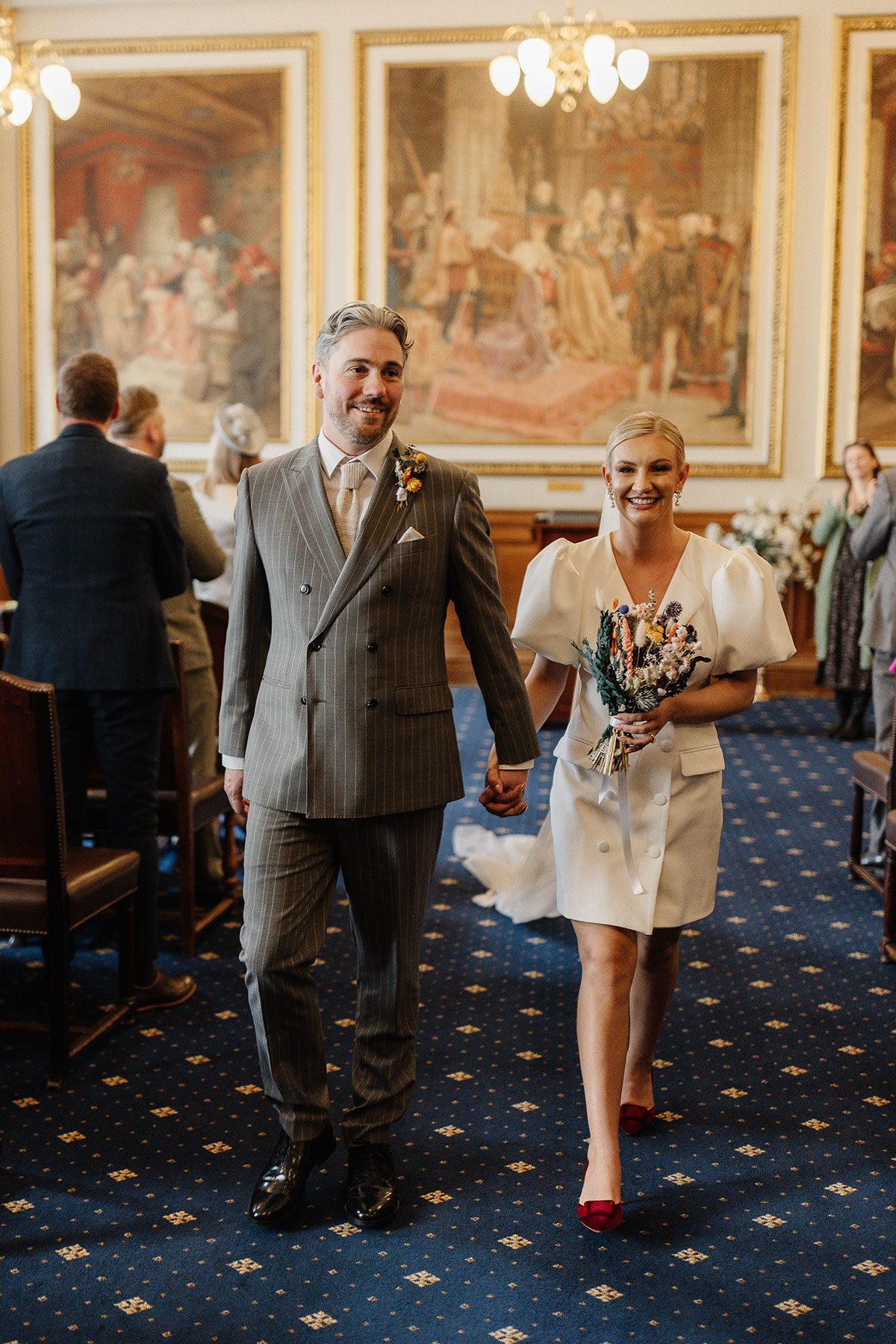 Newlyweds walk hand-in-hand down the aisle at Edinburgh City Chambers after their registry office ceremony, the bride wearing a short puff-sleeve dress and red heels.