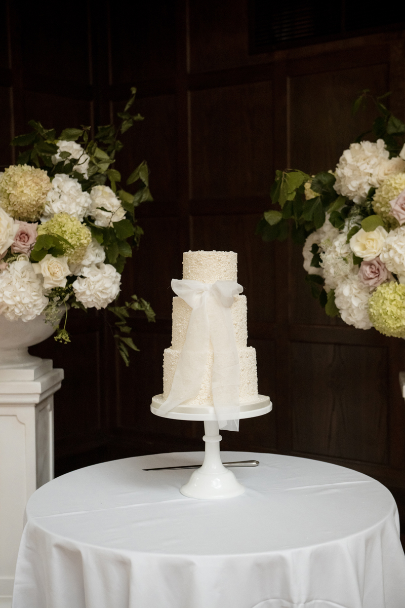 Three-tier white wedding cake with bow detail surrounded by white and blush flowers at Crossbasket Castle near Glasgow.