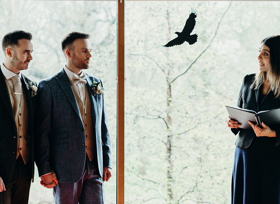 two grooms holding hands during their wedding ceremony while looking at a female officiant carrying a binder on right side of picture