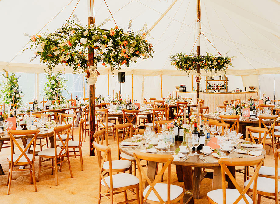 A marquee set for a wedding reception dinner with circular tables and wooden cross back chairs. Circular flower decorations hang from the ceiling, there is a white-wooden-clad bar in the background and all the tables are filled with wine glasses, wine bottles, flower decorations and candles