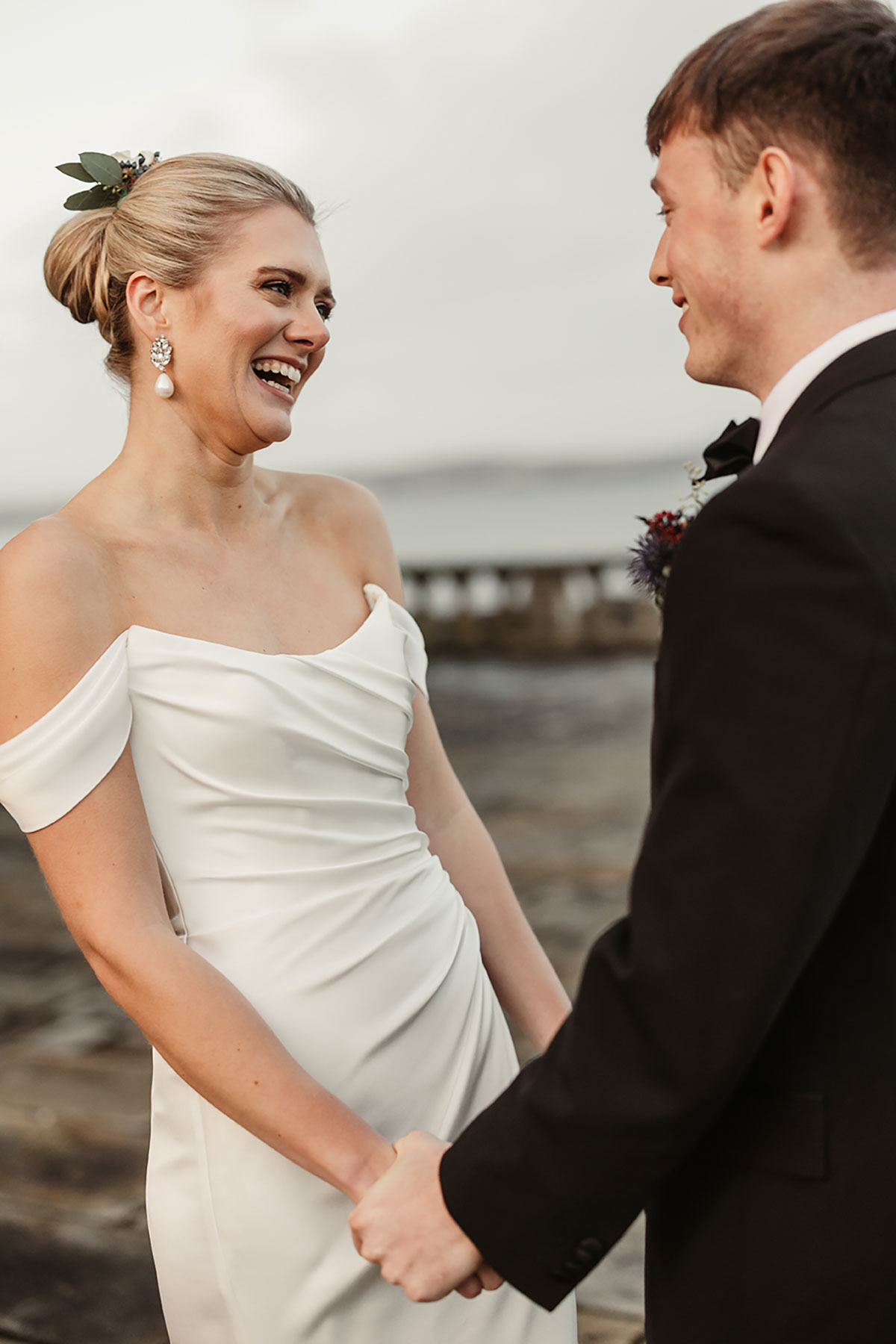 Bride laughing while holding hands with the groom outdoors, with water and soft winter light in the background.