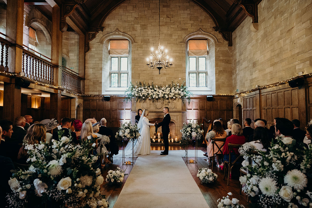 Wedding ceremony in Achnagairn Castle's historic ballroom with floral aisle arrangements, candlelight and guests gathered to witness the vows