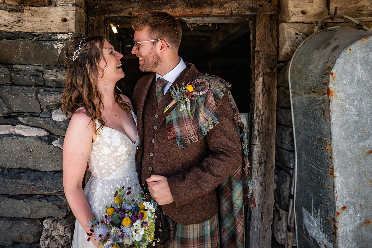 Bride and groom laugh together beside a rustic stone building doorway. The bride wears a lace wedding dress and holds a wildflower bouquet, while the groom wears a brown tweed jacket with tartan kilt, fly plaid and boutonniere