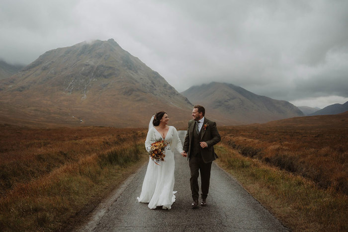 a bride and groom walking hand in hand along a road surrounded by Glencoe mountains