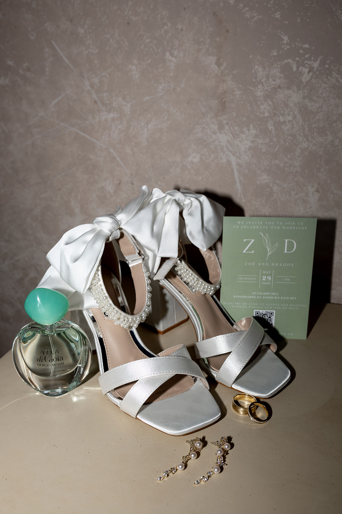 Bridal details including white satin heels with bows, wedding rings, earrings, perfume and an invitation.