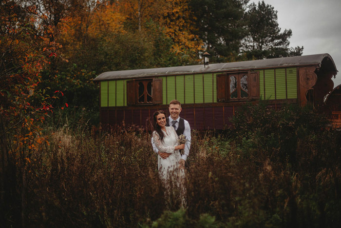  A bride and groom standing in tall grass in front of a green wooden wagon at Roulotte Retreat.