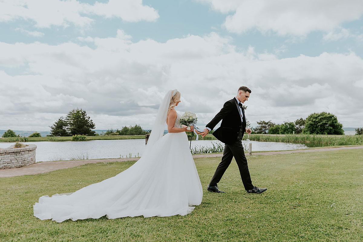 groom in tuxedo walks in front, holding hand behind him to blonde bride in ballgown and long trailing veil as they walk across the grass