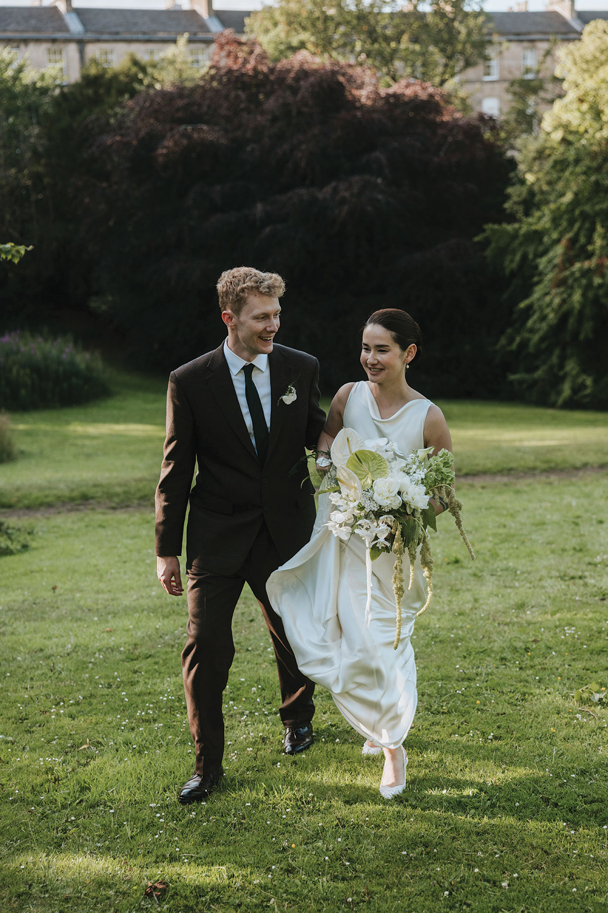 Bride and groom walking across a garden lawn, smiling together on their wedding day.