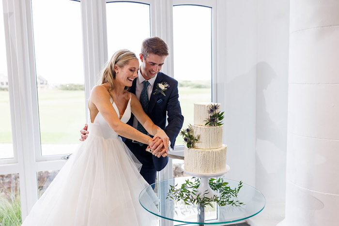 A joyful couple in wedding attire, surrounded by the natural light by a window at the Old Course Hotel, cutting a multi-tiered cake together