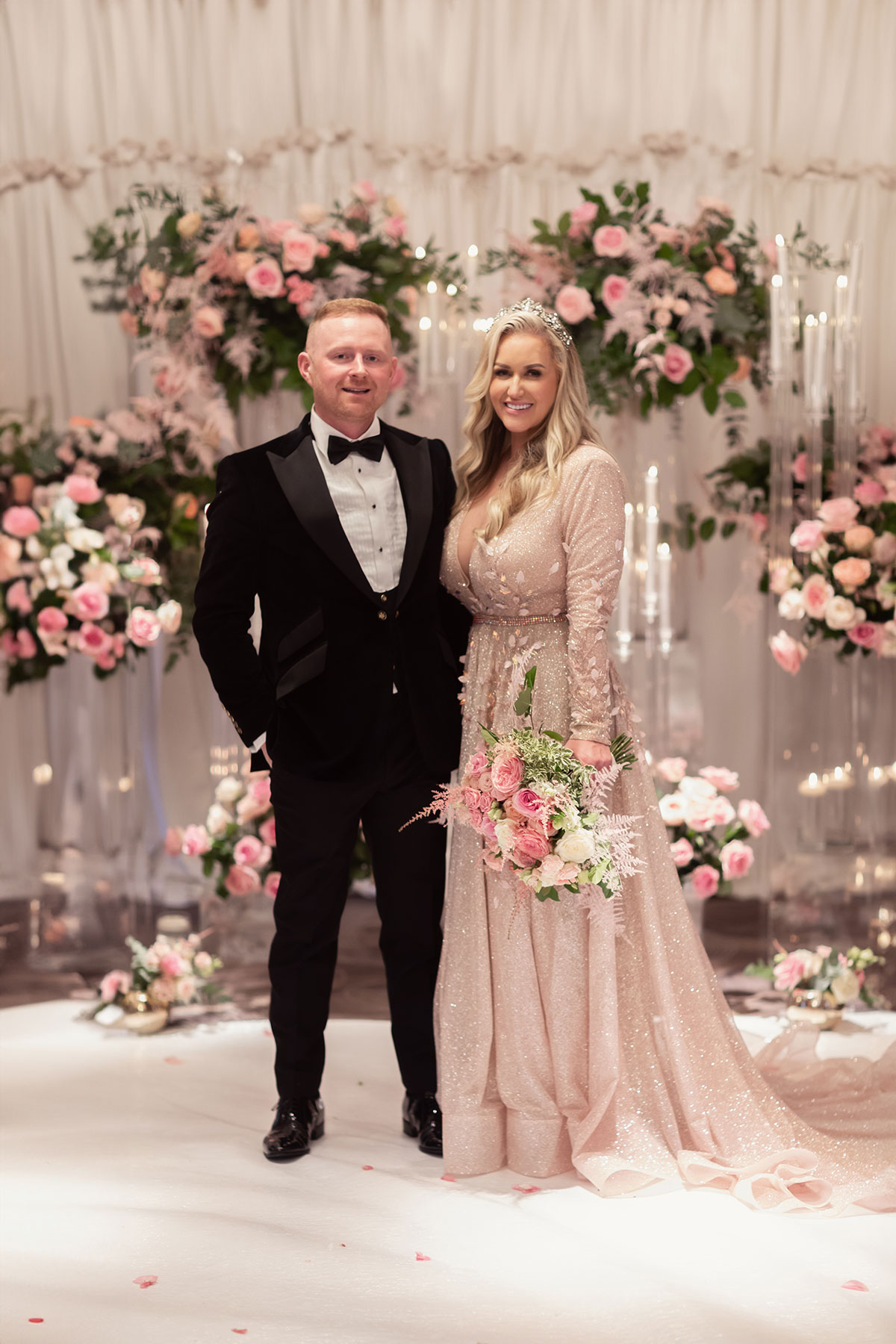 Bride and groom standing beneath floral ceremony arch with pink roses and candle styling