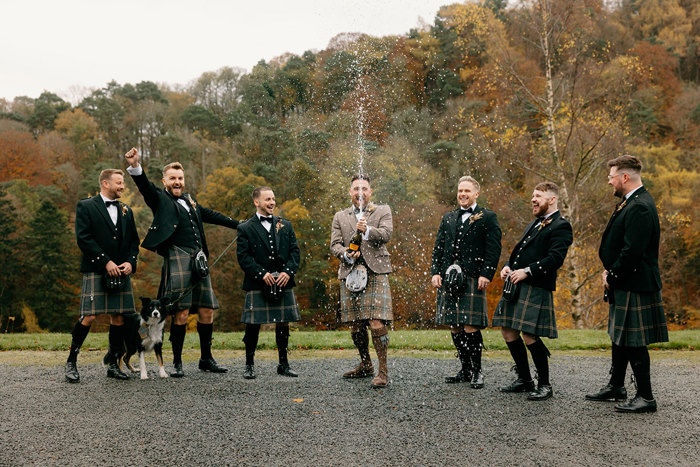 a row of six men wearing kilts standing in front of trees. One of them is wearing a lighter coloured kilt outfit and spraying from a bottle of champagne