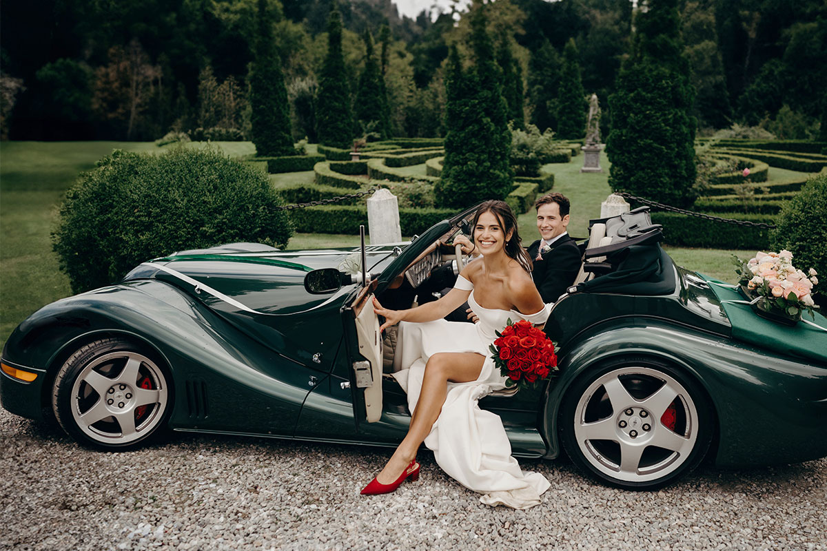 A bride stepping out or a green convertible car holding a red bouquet and wearing red shoes, a groom sits in the driver's seat smiling at the camera