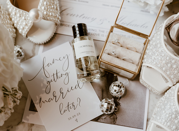 a wedding details flat lay with wedding invitation, bottle of perfume, wedding and engagement rings in gold-frame glass box and pearl covered wedding shoes