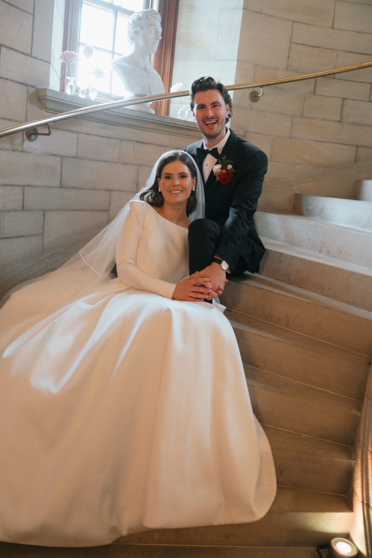 Nicole and Ryan pose together on the stone staircase inside Barnbougle Castle, with Nicole's Suzanne Neville wedding dress spread across the steps.