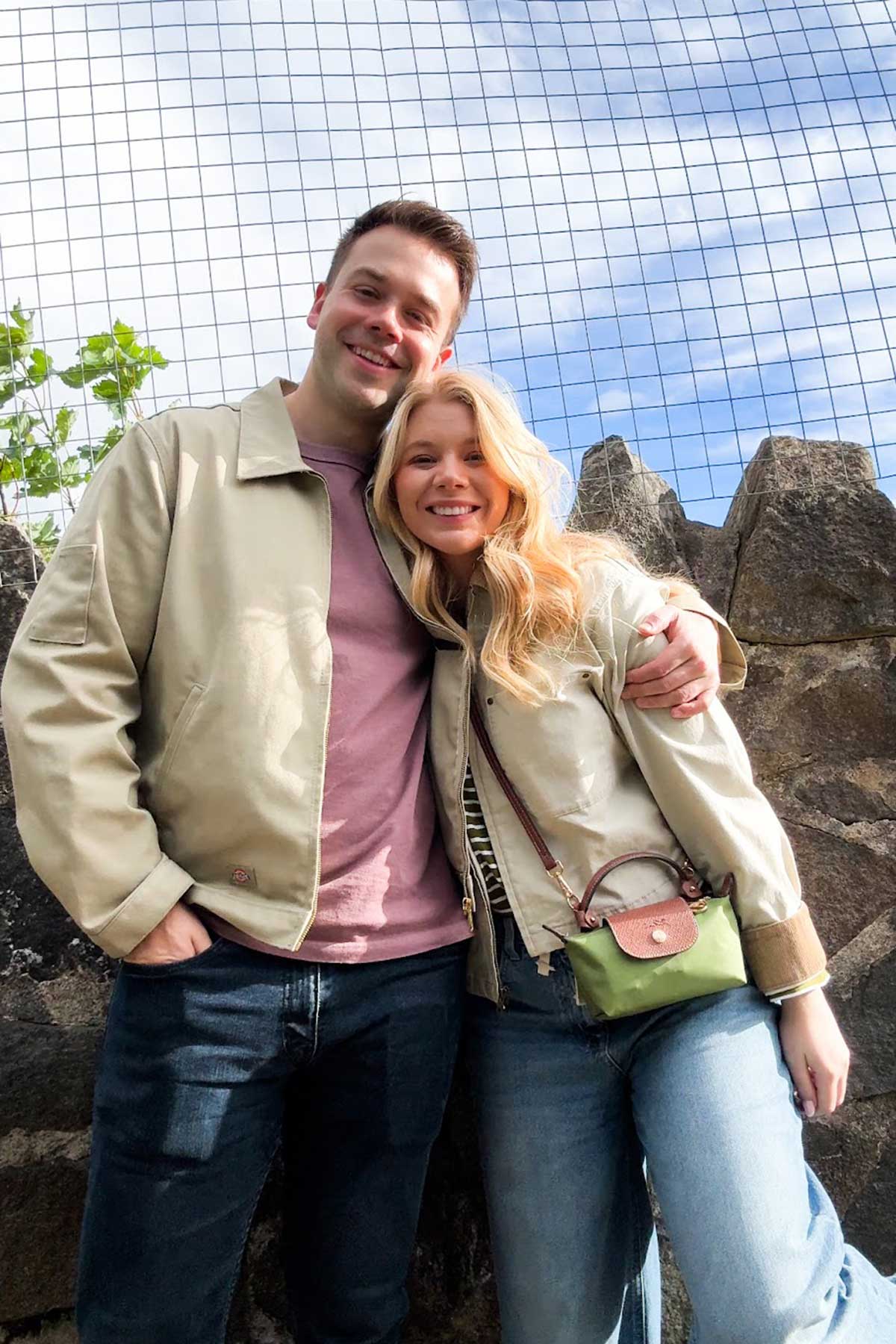 Couple standing arm in arm outdoors against a stone wall and blue sky, smiling at the camera