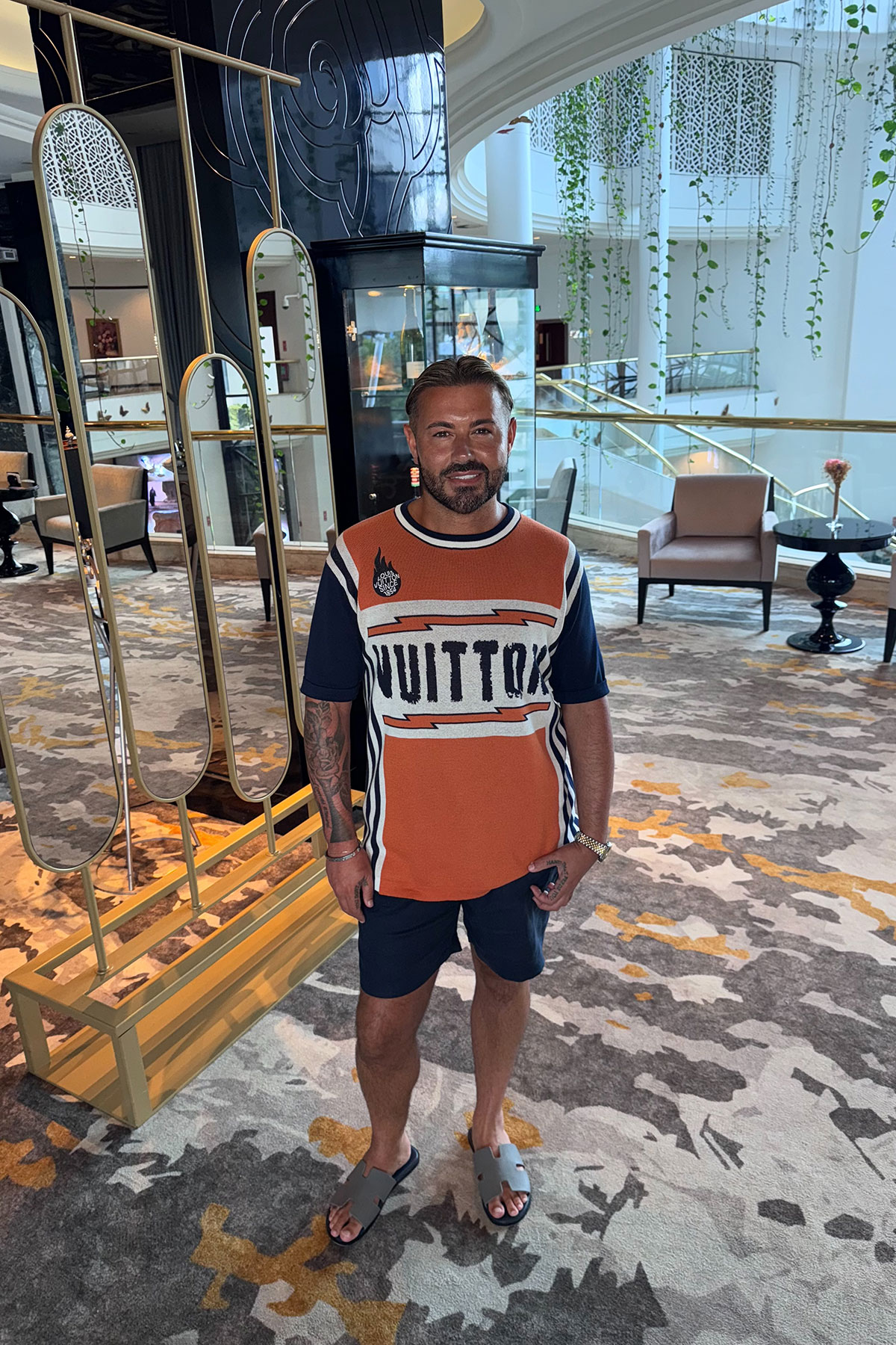 Man standing in a modern hotel lobby, wearing an orange and navy Louis Vuitton T-shirt, navy shorts and grey sliders, smiling at the camera