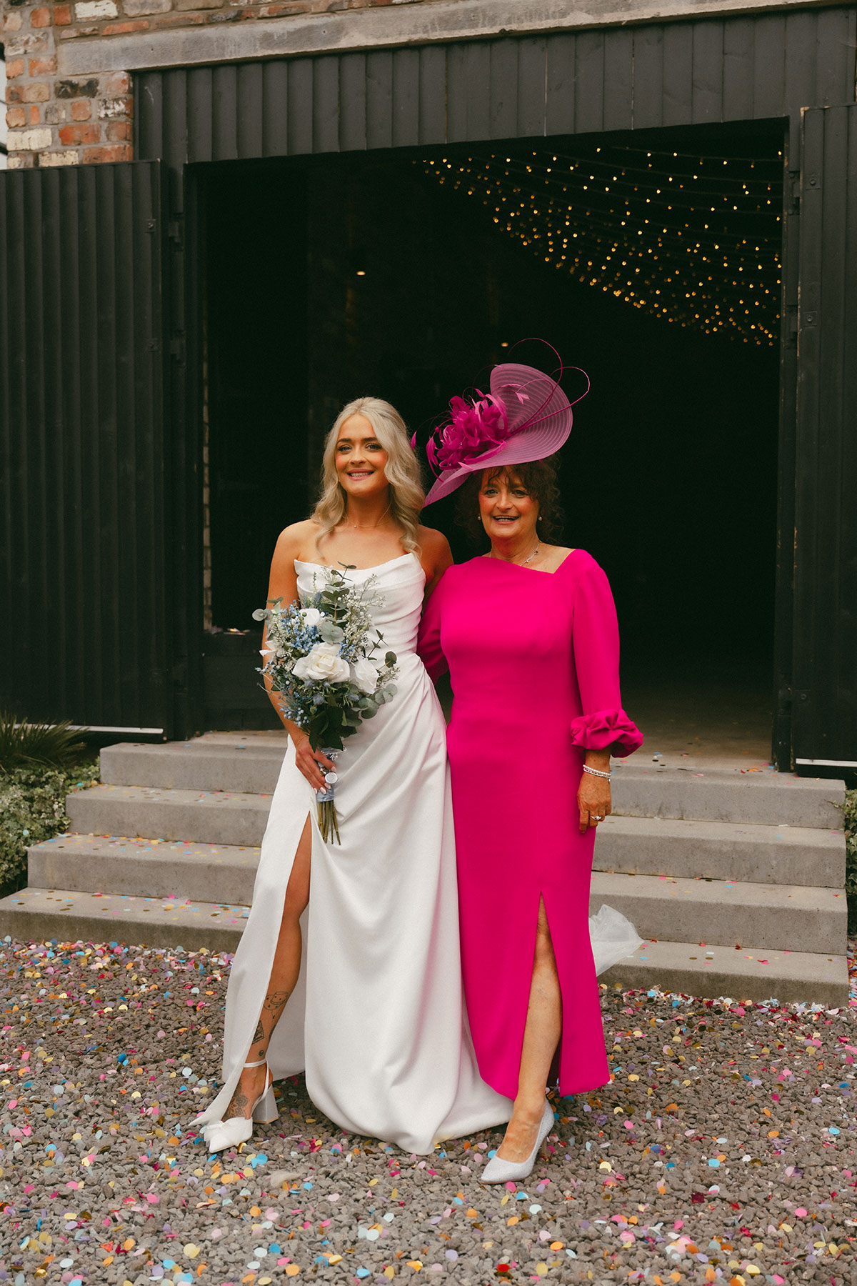 Bride in a white gown holding a bouquet stands beside an older woman in a bright pink dress and fascinator outside a barn, with confetti scattered on the ground