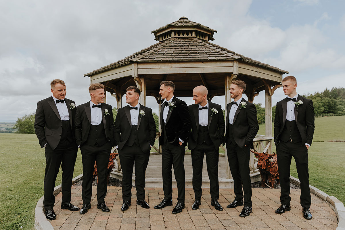 seven men in black tuxedos and shiny shoes stand in a line, looking at one another, in front of a bandstand surrounded by grass