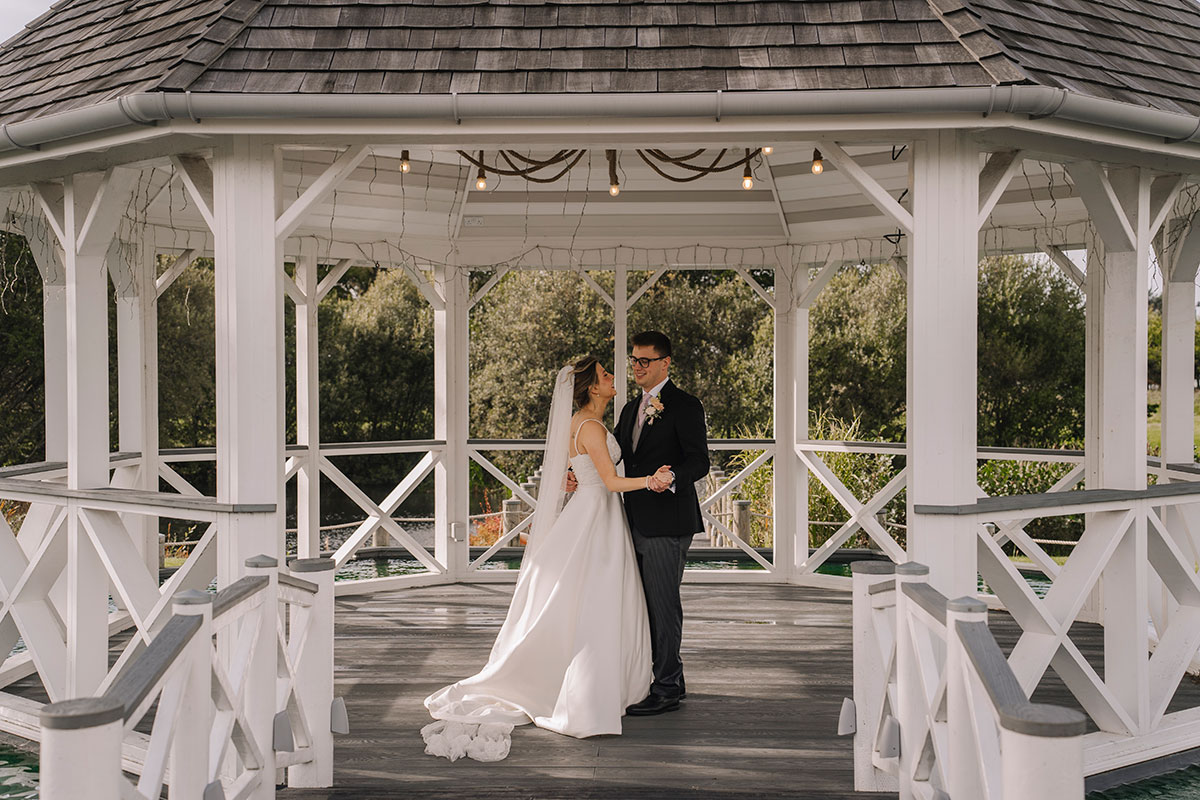 Newlywed couple sharing a quiet moment together beneath a white gazebo in the venue gardens
