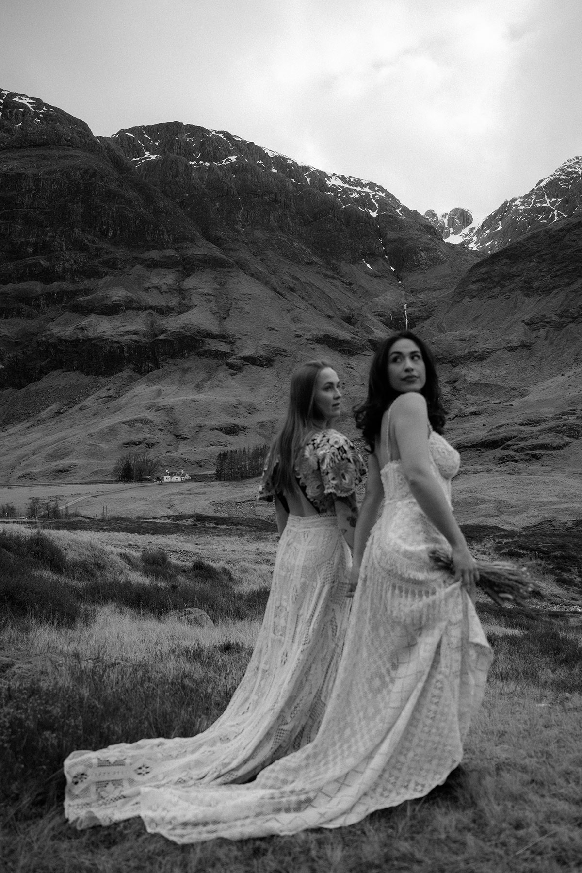 Black and white image of a couple in lace wedding dresses looking across the Glencoe landscape during a Scottish elopement shoot.
