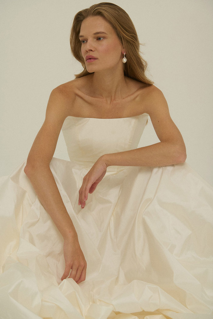close up on a woman wearing a pearl earrings and a strapless corset ballgown wedding dress sat down with the dress gathered out around her, her arms resting on her knees on top of her dress