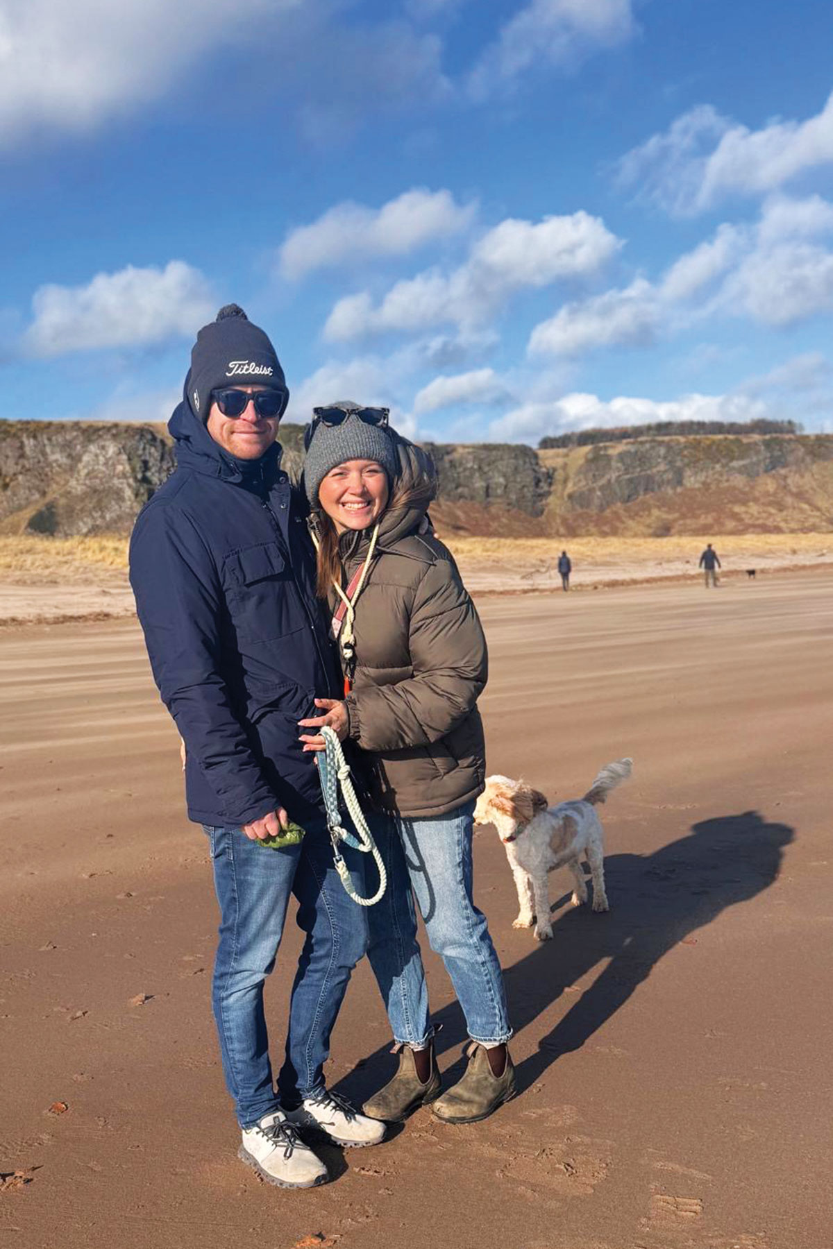 Couple standing on a sandy beach with cliffs in the background; both wear winter jackets and hats, with a small dog on a lead beside them