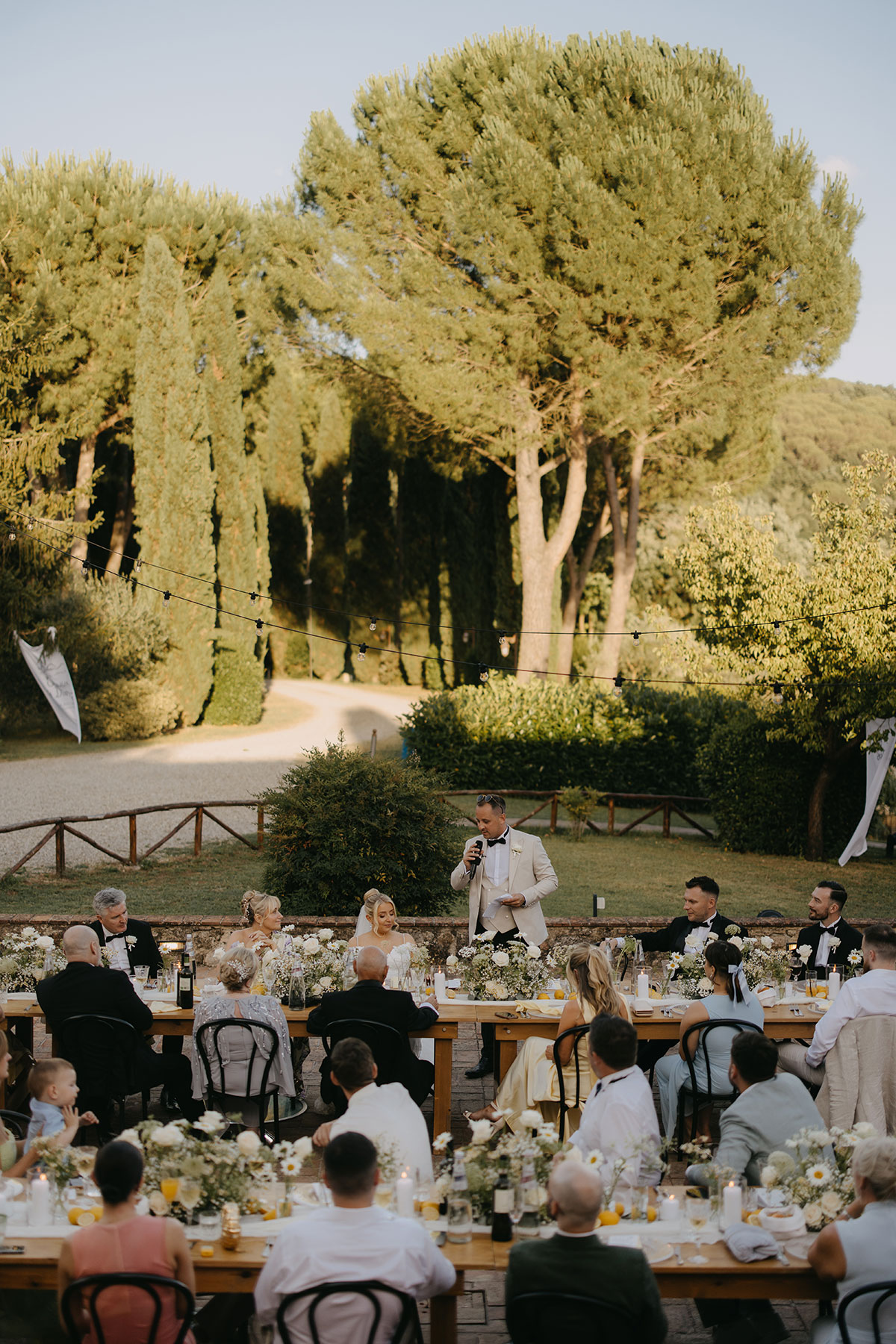 Guests seated at long wooden tables during an outdoor wedding reception surrounded by trees and greenery.
