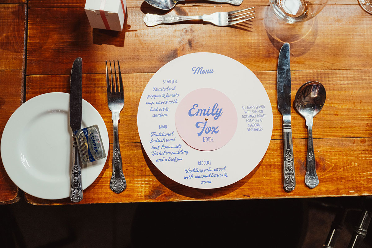 Wedding place setting with personalised circular menu reading “Emily Fox”, surrounded by cutlery, a bread plate, wine glasses and a wooden banquet table surface.