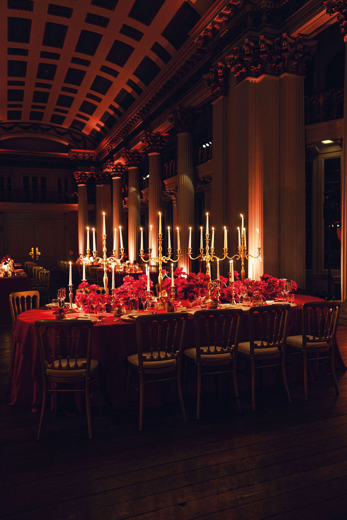 Long reception table styled with red florals, gold candelabras and red table linens beneath warm ambient lighting in a grand historic venue