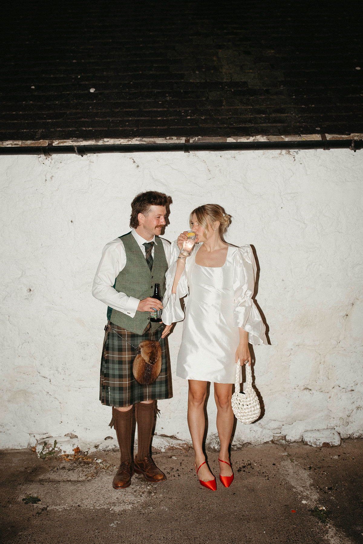 Bride in short reception dress and groom in kilt pose for relaxed nighttime portrait after Highland barn wedding