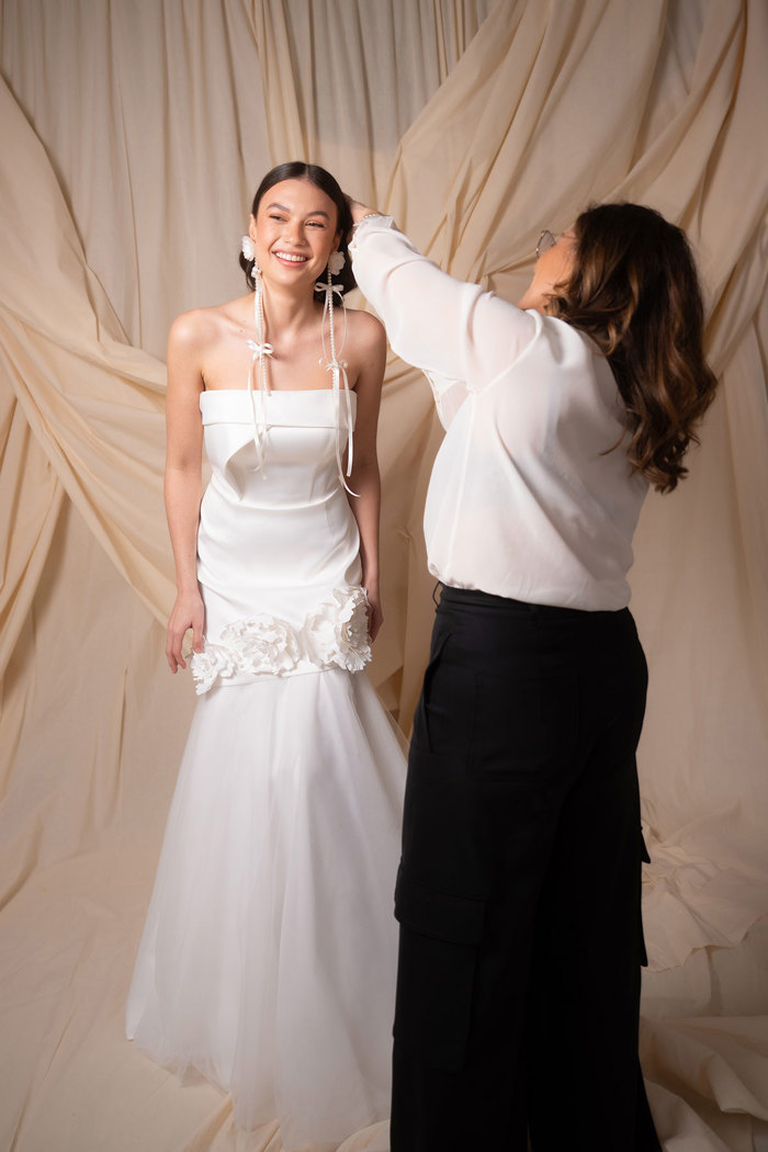 dark haired model wears strapless wedding dress and very long white floral and bow dangly earrings and smiles as a woman in all black adjusts her hair