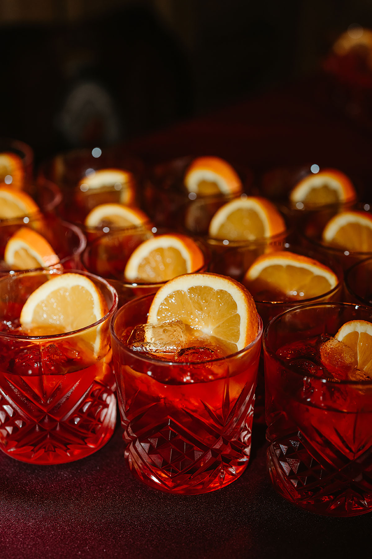 Rows of red Negroni cocktails served in cut-glass tumblers, each garnished with a slice of orange.
