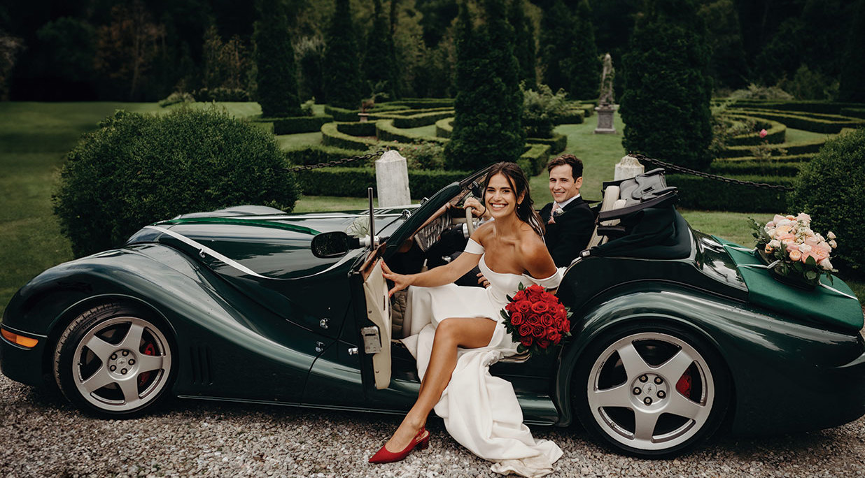 a bride stepping out of an open-top racing green sports car with the groom in the driving seat behind her. She is carrying a bouquet of red roses. The gardens of Achnagairn Castle are in the background.