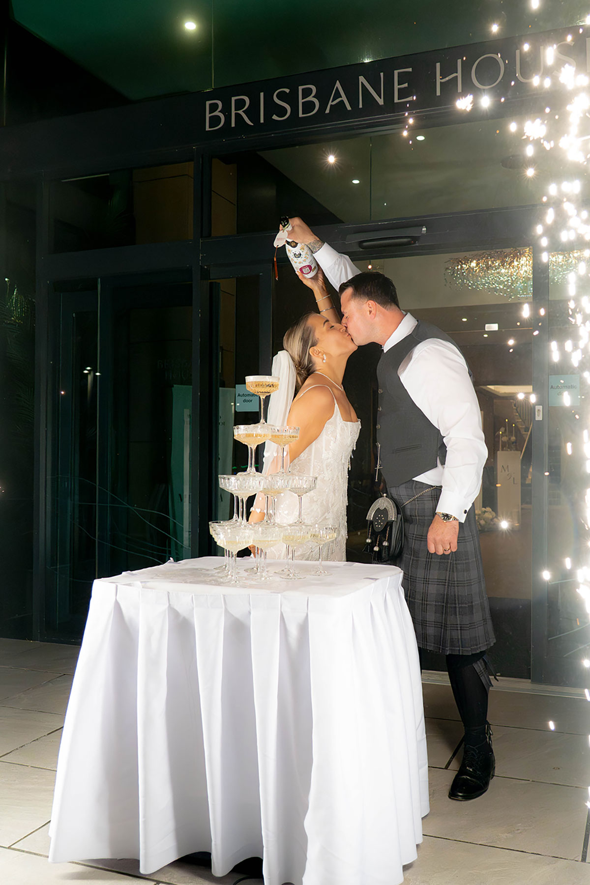Bride and groom kissing beside champagne tower and sparklers outside Brisbane House Hotel evening reception