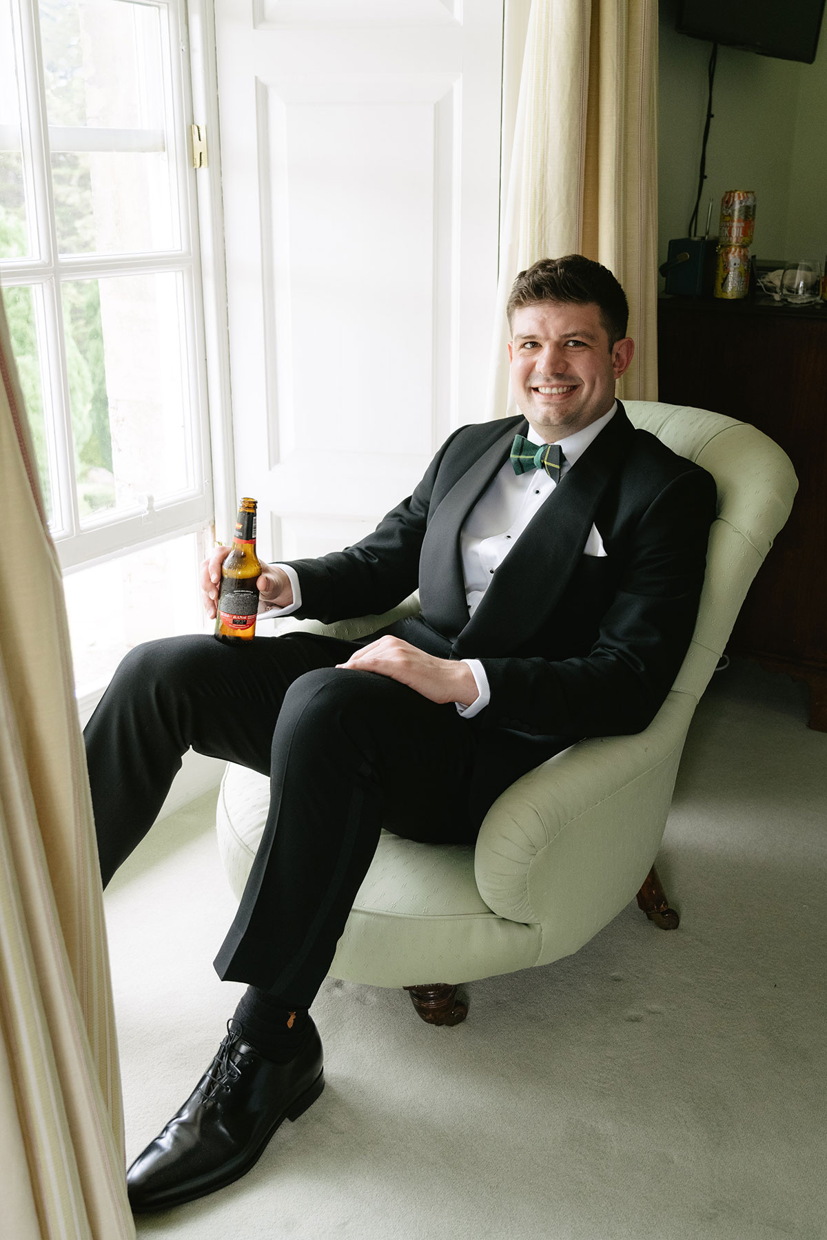 Groom in black tuxedo sitting with drink during wedding morning preparations at Gilmerton House
