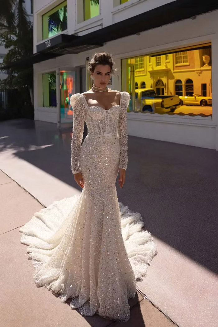 brunette woman wears sparkly bold shouldered long sleeve mermaid style wedding dress out on the pavement in front of brightly coloured window storefront