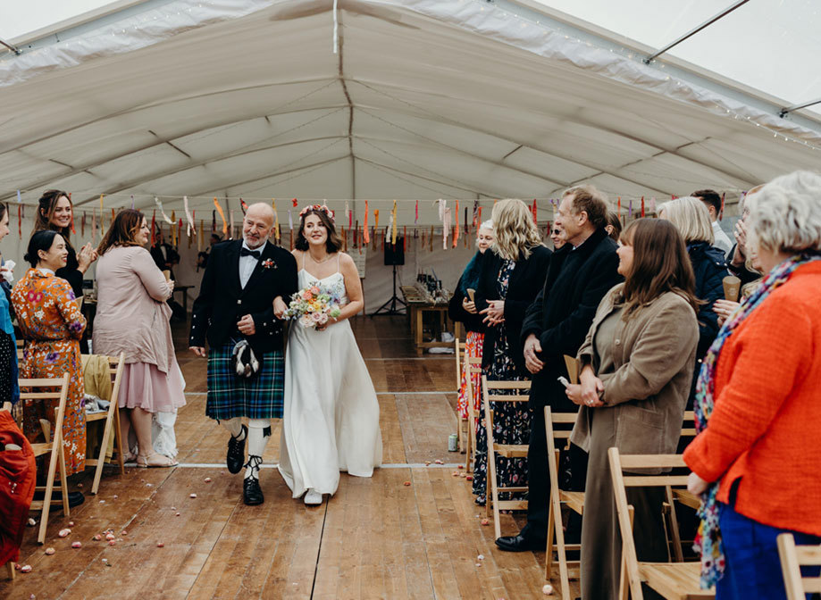 a bride wearing a flower crown walks down the aisle on the arm of a man wearing a kilt. Wedding guests standing on either side turn to look at them. The marquee setting has a wooden floor and colourful paper streamers on strings hanging in the background