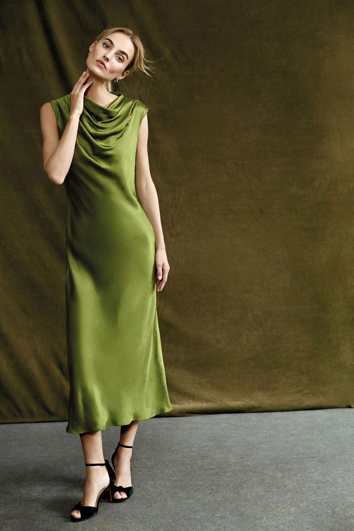 Olive green satin midi dress with draped cowl neckline styled against a textured studio backdrop